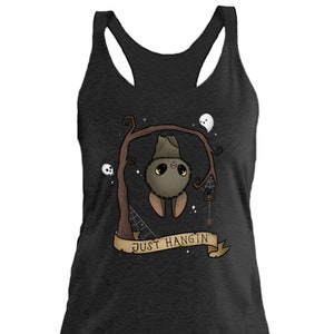 May include: A dark gray racerback tank top with a cute cartoon bat hanging from a tree branch. The bat has big eyes and a friendly smile. The text "Just Hangin'" is written on a banner below the bat.