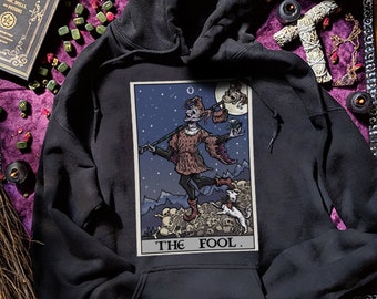 May include: Black hooded sweatshirt with a tarot card design featuring the Fool card. The card depicts a jester figure walking towards a cliff with a dog at his feet. The text "THE FOOL." is printed below the card.