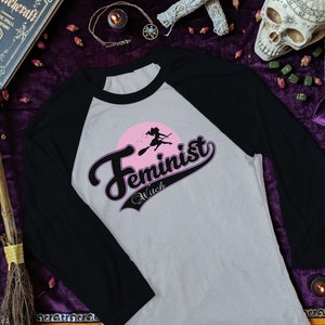 May include: A grey and black baseball tee shirt with a pink and black graphic that says "Feminist Witch" with a silhouette of a witch flying on a broomstick.