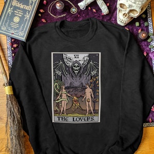 Puede incluir: Sudadera negra con un gr&aacute;fico de la carta del tarot de Los Enamorados. La carta presenta una figura esquel&eacute;tica con una t&uacute;nica y alas extendidas, y dos figuras desnudas de pie a cada lado de un volc&aacute;n. El n&uacute;mero de la carta, VI, est&aacute; en la esquina superior derecha. El texto "THE LOVERS" est&aacute; impreso debajo de la carta.