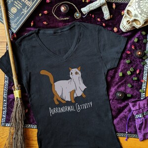 May include: Black v-neck t-shirt with a graphic of a cat wearing a ghost sheet. The text "Purranormal Cativity" is printed below the cat.