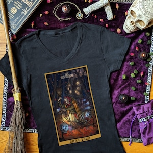 May include: Black v-neck t-shirt with a colorful illustration of Baba Yaga, a witch from Slavic folklore, holding a lantern and a broom. The illustration is in the style of a tarot card, with the text "IX THE HERMIT" and "BABA YAGA" at the top and bottom of the card.