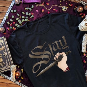 Puede incluir: Camiseta negra con un gr&aacute;fico marr&oacute;n de una mano sosteniendo una daga y la palabra "Slay" en una fuente g&oacute;tica.