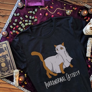 May include: Black t-shirt with a graphic of a cat wearing a ghost sheet. The text "Purranormal Cativity" is printed below the cat.