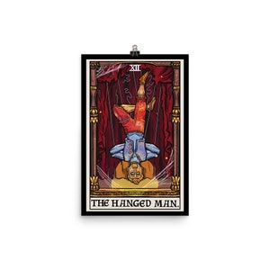 Op de afbeelding: Een tarotkaart-illustratie van De Gehangene. De kaart is ingelijst in een zwarte rand met gouden accenten. De kaart toont een man die ondersteboven aan een touw hangt, met gekruiste benen. De man draagt een blauw shirt, een rode broek en een gele hoed. De achtergrond is een rood gordijn met gouden versieringen.
