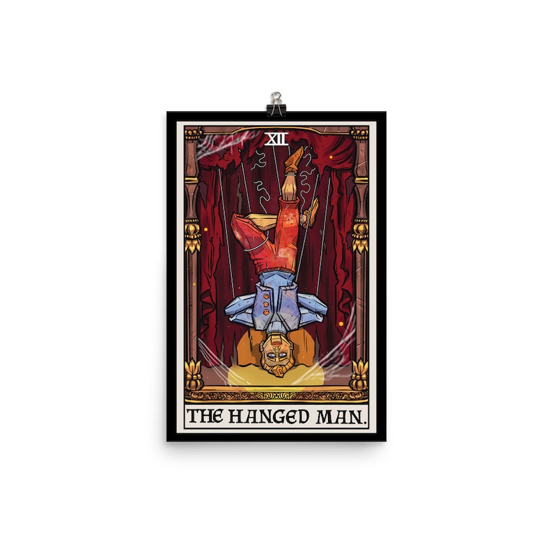 Op de afbeelding: Een tarotkaart-illustratie van De Gehangene. De kaart is ingelijst in een zwarte rand met gouden accenten. De kaart toont een man die ondersteboven aan een touw hangt, met gekruiste benen. De man draagt een blauw shirt, een rode broek en een gele hoed. De achtergrond is een rood gordijn met gouden versieringen.