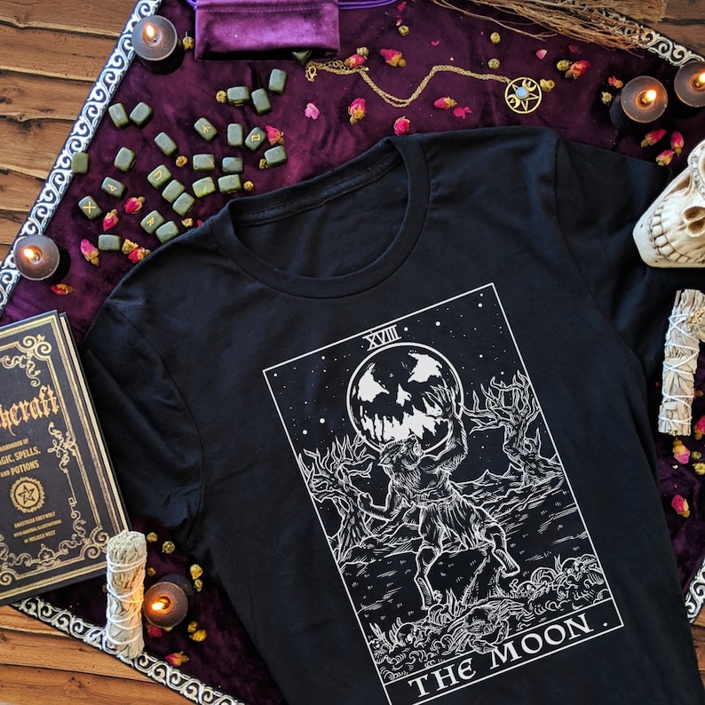 May include: Black t-shirt featuring a white graphic of The Moon tarot card. The card depicts a werewolf-like creature holding a crescent moon with a face. The text "XVIII THE MOON" is printed at the bottom of the card.