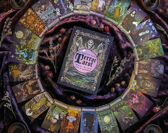 Könnte beinhalten: Ein Tarot-Deck und ein Führer, "The Ghoulish Garb Terror Tarot", sind von einzelnen Tarotkarten umgeben, die in einem Kreis angeordnet sind. Die Karten zeigen farbenfrohe Illustrationen mit gotischen und Fantasy-Themen. Das Deck liegt auf einem dunkelvioletten Samtstoff.