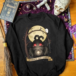 May include: Black sweatshirt with a graphic of a cute black moth holding a lantern. The moth has red eyes and is sitting on a branch under a full moon. The text "Embrace the Light" is written on a banner below the moth.