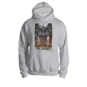 Puede incluir: Una sudadera con capucha gris con una impresi&oacute;n gr&aacute;fica de una carta de tarot que representa a Los Amantes. La carta presenta una figura esquel&eacute;tica con alas extendidas, dos figuras desnudas y un volc&aacute;n.