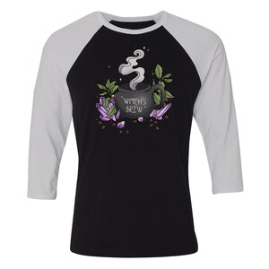 Op de afbeelding: Een zwart en wit baseballshirt met een zwarte koffiemok met de tekst "Witch's Brew" omringd door paarse kristallen en groene bladeren.