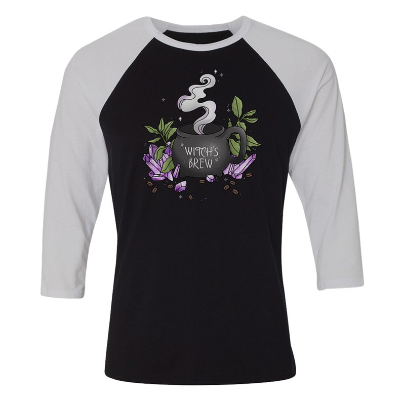 Op de afbeelding: Een zwart en wit baseballshirt met een zwarte koffiemok met de tekst "Witch's Brew" omringd door paarse kristallen en groene bladeren.