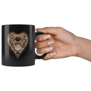 Puede incluir: Taza de cer&aacute;mica negra con un dise&ntilde;o en forma de coraz&oacute;n con una ilustraci&oacute;n de ojo, sol y luna. La taza tiene un asa y est&aacute; sujeta por una mano. El dise&ntilde;o es en tonos marrones y beige.