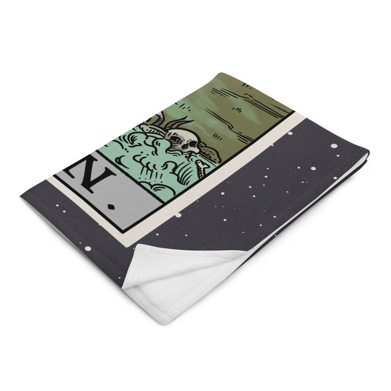 May include: A black and white throw blanket with a tarot card design featuring a skeleton in a green and brown landscape. The card reads "Death".