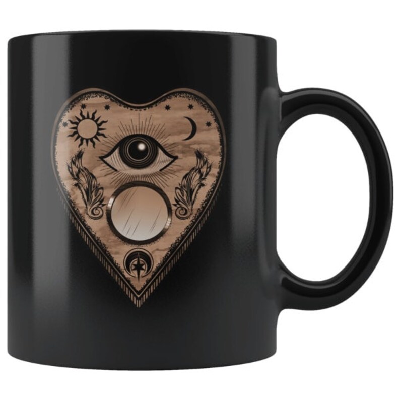 Puede incluir: Taza de cer&aacute;mica negra con un dise&ntilde;o en forma de coraz&oacute;n con elementos celestiales y m&iacute;sticos. El dise&ntilde;o incluye un ojo, sol, luna, plumas y un c&iacute;rculo espejado. La taza tiene un asa negra y un acabado brillante.