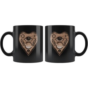 Puede incluir: Dos tazas de cer&aacute;mica negra con un dise&ntilde;o en forma de coraz&oacute;n. El dise&ntilde;o presenta un ojo, un sol, una luna y otros s&iacute;mbolos con un acabado de madera. Las tazas son id&eacute;nticas y aptas para bebidas fr&iacute;as o calientes.