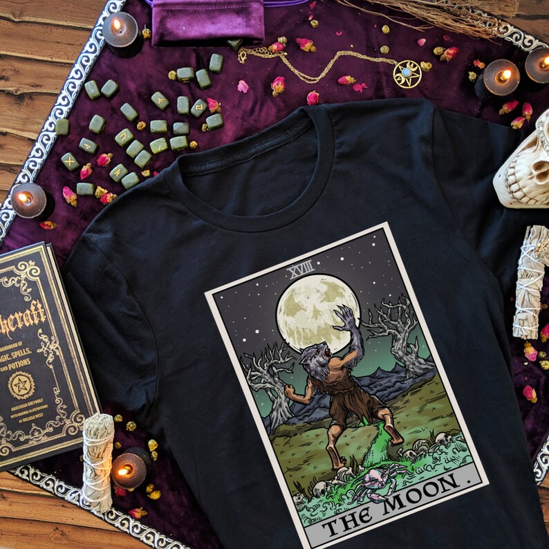Peut inclure: T-shirt noir avec un graphique de la carte de tarot La Lune. La carte repr&eacute;sente un loup-garou hurlant &agrave; la lune, avec un fond d'arbres et d'&eacute;toiles. Le num&eacute;ro de la carte, XVIII, est dans le coin sup&eacute;rieur gauche.