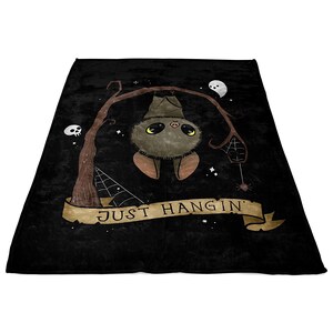 Pu&ograve; includere: Una coperta in pile nero con un simpatico pipistrello dei cartoni animati appeso a un ramo. Il pipistrello ha la pelliccia marrone e occhi grandi. Il testo "JUST HANG IN'" &egrave; scritto su uno striscione sotto il pipistrello.