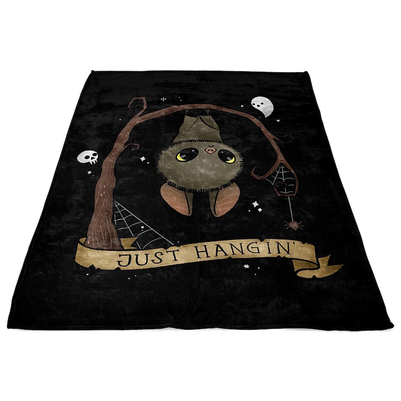 Pu&ograve; includere: Una coperta in pile nero con un simpatico pipistrello dei cartoni animati appeso a un ramo. Il pipistrello ha la pelliccia marrone e occhi grandi. Il testo "JUST HANG IN'" &egrave; scritto su uno striscione sotto il pipistrello.