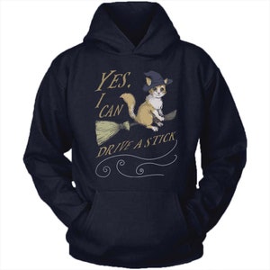 Pode incluir: Um moletom azul marinho com capuz com um gr&aacute;fico de um gato usando um chap&eacute;u de bruxa e montando uma vassoura. O texto no moletom diz "YES, I CAN DRIVE A STICK."