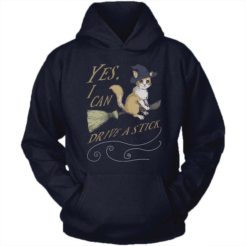 Pode incluir: Um moletom azul marinho com capuz com um gr&aacute;fico de um gato usando um chap&eacute;u de bruxa e montando uma vassoura. O texto no moletom diz "YES, I CAN DRIVE A STICK."
