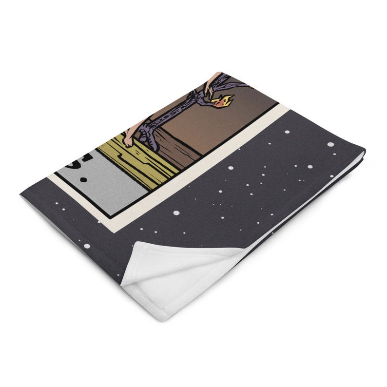 May include: A gray fleece throw blanket with a white underside. The blanket features a colorful illustration of a tarot card with a person climbing a tree. The card is surrounded by a black border and a white background with black dots.