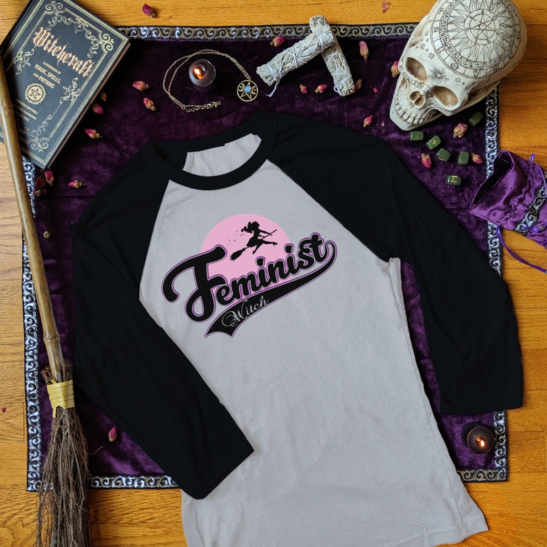 May include: A grey and black baseball tee shirt with a pink and black graphic that says "Feminist Witch" with a silhouette of a witch flying on a broomstick.