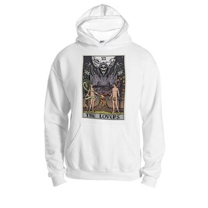 Puede incluir: Sudadera con capucha blanca con un estampado gr&aacute;fico de una carta del tarot que representa la carta de los Enamorados. La carta muestra una figura esquel&eacute;tica con alas, dos figuras de pie debajo, y el texto "THE LOVERS".