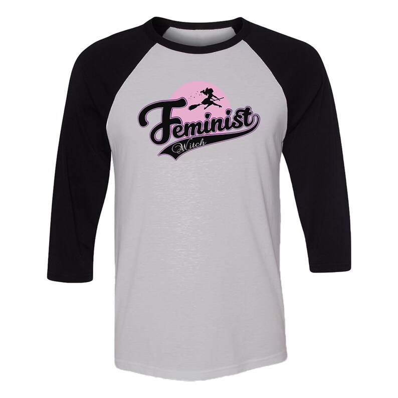 May include: A white and black baseball style t-shirt with a pink and black graphic that says "Feminist Witch" with a silhouette of a witch flying on a broomstick.