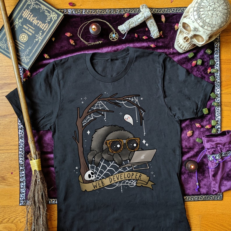 May include: Black t-shirt featuring a cartoon spider wearing glasses and working on a laptop. The design includes a spider web, a ghost, and the text "WEB DEVELOPER" on a banner. The shirt is surrounded by witchy items.