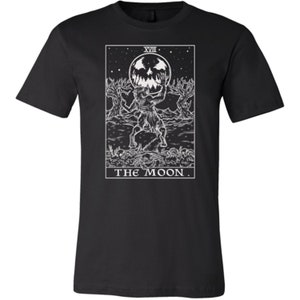 May include: Black and white graphic t-shirt featuring a tarot card design. The card depicts the "The Moon" card with a werewolf-like creature holding a crescent moon with a face. The card number "XVIII" is at the top of the card.