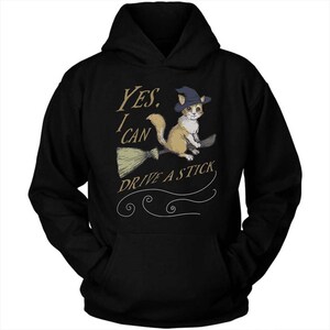 Pode incluir: Camisola de capuz preta com um gr&aacute;fico de um gato a usar um chap&eacute;u de bruxa e a andar de vassoura. O texto diz "YES, I CAN DRIVE A STICK."