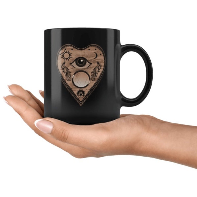 Puede incluir: Una taza de cer&aacute;mica negra con un dise&ntilde;o en forma de coraz&oacute;n con s&iacute;mbolos celestiales y espirituales. El dise&ntilde;o incluye un sol, una luna, un ojo y otros elementos decorativos. La taza se sostiene en una mano.