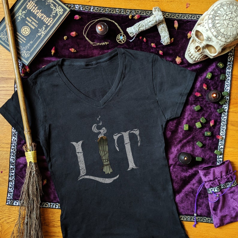 May include: Black V-neck t-shirt with the word "LIT" in gray, featuring a graphic of a burning sage bundle. The shirt is surrounded by witchy items, including a broom, a book, a skull, and a purple velvet cloth.