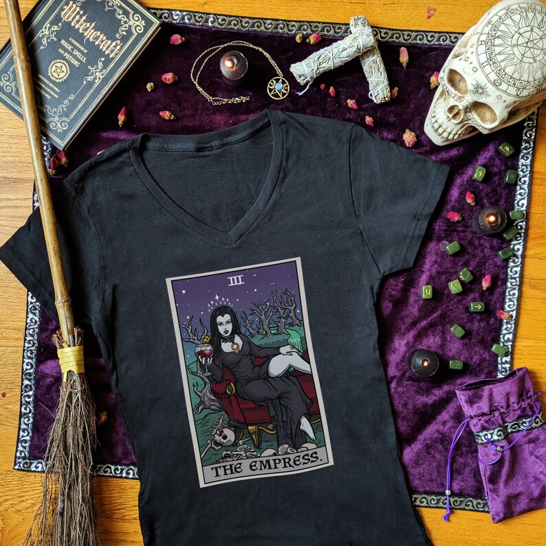 May include: Black v-neck t-shirt with a graphic of the Empress tarot card. The card features a woman in a black dress sitting on a throne with a glass of wine in her hand. The card is surrounded by a purple and black background with stars and trees.