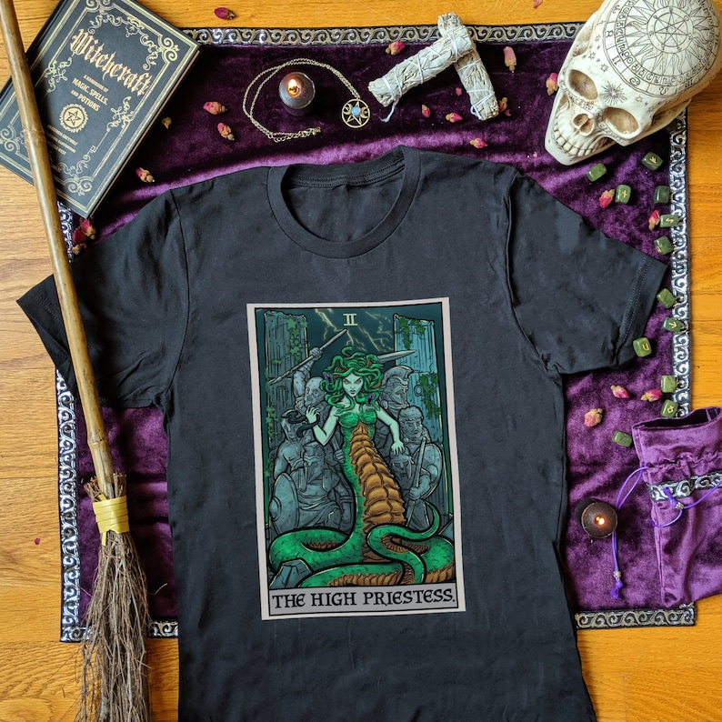May include: Black t-shirt with a graphic print of the High Priestess tarot card featuring a green snake and a woman with snakes for hair. The card is surrounded by a black border and the text "The High Priestess" is printed below the card.