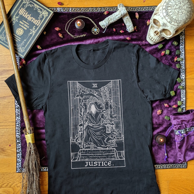 Op de afbeelding: Zwart T-shirt met een witte afbeelding van de Justice-kaart uit een tarotdeck. De kaart toont een figuur in een gewaad die een weegschaal en een zwaard vasthoudt, met het Romeinse cijfer XI en het woord "JUSTICE" eronder.
