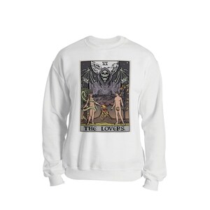Puede incluir: Sudadera blanca con una ilustraci&oacute;n en blanco y negro de una carta del tarot que representa a Los Enamorados. La carta muestra una figura esquel&eacute;tica con alas y una t&uacute;nica, de pie sobre dos figuras desnudas. La carta est&aacute; etiquetada como "VI LOS ENAMORADOS."