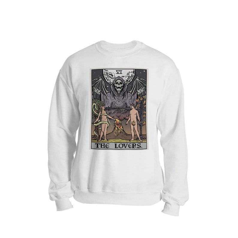 Puede incluir: Sudadera blanca con una ilustraci&oacute;n en blanco y negro de una carta del tarot que representa a Los Enamorados. La carta muestra una figura esquel&eacute;tica con alas y una t&uacute;nica, de pie sobre dos figuras desnudas. La carta est&aacute; etiquetada como "VI LOS ENAMORADOS."