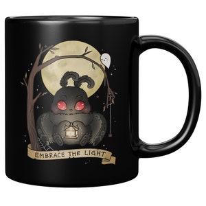May include: Black ceramic mug with a graphic of a black moth holding a lantern. The moth has red eyes and is sitting in front of a full moon. The text "Embrace the Light" is written on a banner below the moth.