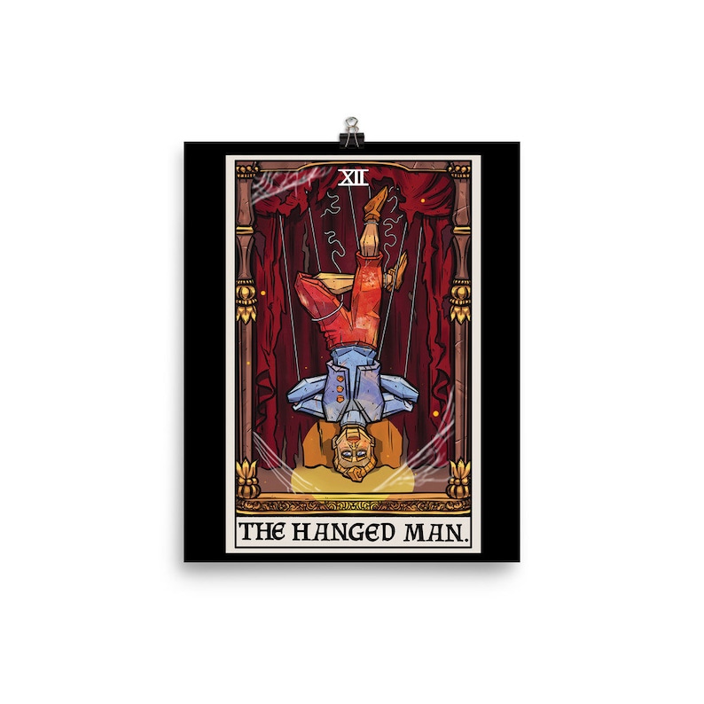Op de afbeelding: Een tarotkaart-illustratie van De Gehangene. De kaart is zwart met een gouden lijst en toont een man die ondersteboven aan een tak hangt. De man draagt een blauw shirt en een rode broek. De kaart is getiteld "De Gehangene" in gouden letters.
