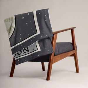 May include: A gray fleece throw blanket with a white border and a moon graphic. The blanket has white dots on it and the text "MOON." in black.