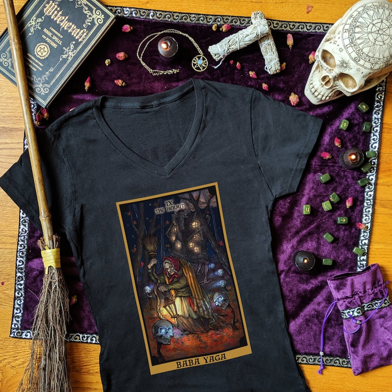 May include: Black v-neck t-shirt with a colorful illustration of Baba Yaga, a witch from Slavic folklore, holding a lantern and a broom. The illustration is in the style of a tarot card, with the text "IX THE HERMIT" and "BABA YAGA" at the top and bottom of the card.