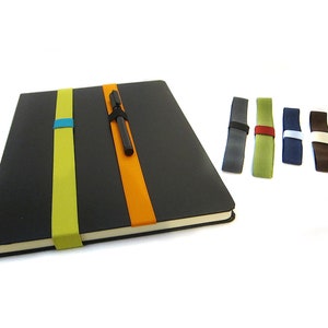 May include: A black notebook with a pen holder and three elastic bands in different colours. The notebook has a green, orange, and blue elastic band. There are five additional elastic bands in different colours: grey, green, blue, brown, and white.