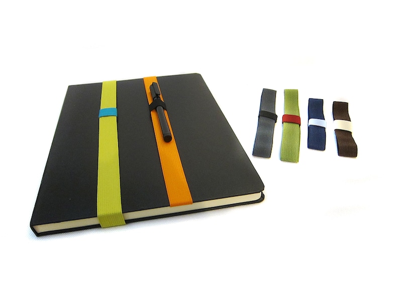 May include: A black notebook with a pen holder and three elastic bands in different colours. The notebook has a green, orange, and blue elastic band. There are five additional elastic bands in different colours: grey, green, blue, brown, and white.