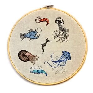 May include: Embroidered hoop art featuring various sea creatures, including a blue anglerfish, a brown nautilus, a red shrimp, a blue jellyfish, a black jellyfish, and a seahorse.