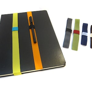 May include: A black notebook with a yellow and orange elastic pen holder. The notebook has a pen tucked into the holder. Five additional elastic pen holders are shown in different colours: grey, green, blue, white, and brown.