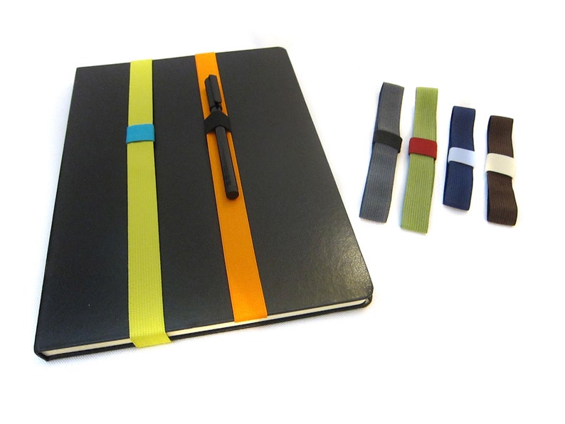 May include: A black notebook with a yellow and orange elastic pen holder. The notebook has a pen tucked into the holder. Five additional elastic pen holders are shown in different colours: grey, green, blue, white, and brown.