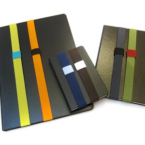 May include: Three different sized notebooks with elastic bands in various colours. The largest notebook is black with yellow, orange, and blue bands. The medium notebook is brown with green, grey, and red bands. The smallest notebook is black with brown, blue, and white bands.