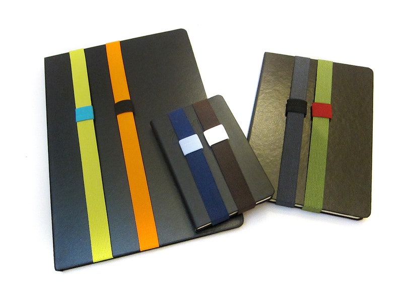May include: Three different sized notebooks with elastic bands in various colours. The largest notebook is black with yellow, orange, and blue bands. The medium notebook is brown with green, grey, and red bands. The smallest notebook is black with brown, blue, and white bands.